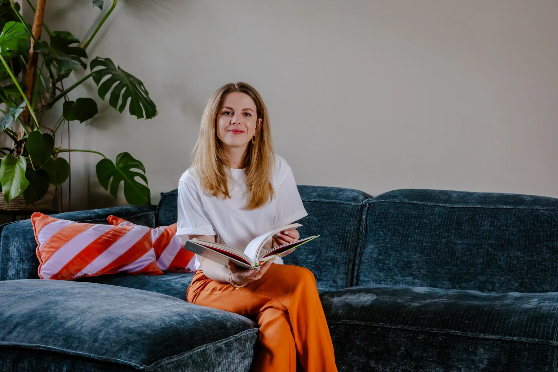 Fleur zit met een boek op de bank en kijkt glimlachend in de camera